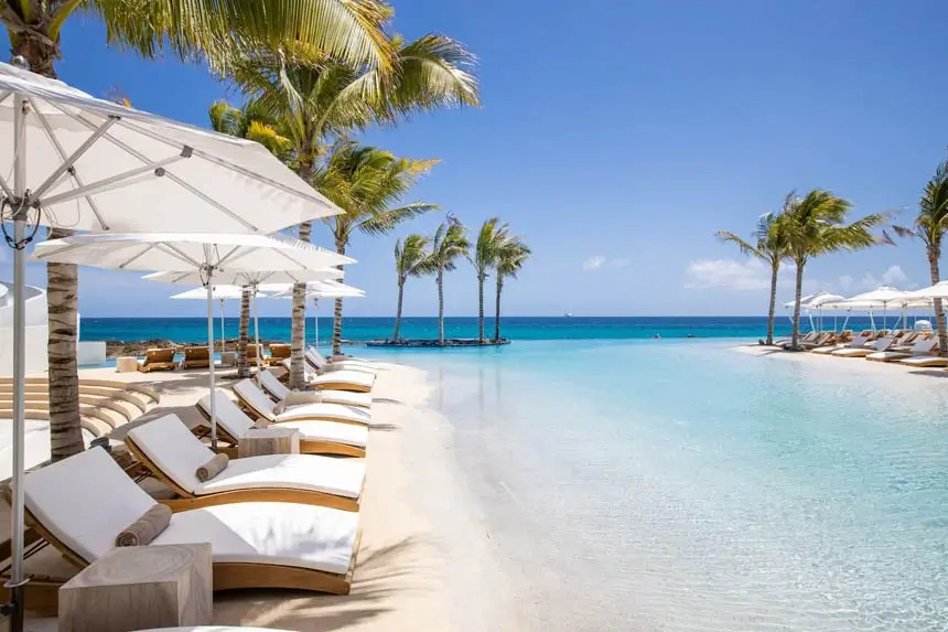 Zero-entry beach-style pool with white sand shoreline, palm trees, and lounge chairs beneath umbrellas